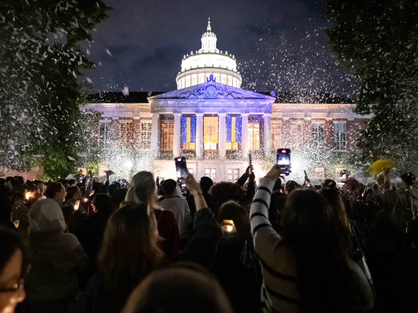 A crowd gathers outside a University of Rochester building, celebrating as colorful confetti falls from the sky.