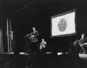 John F. Kennedy giving a speech in an auditorium at a podium, with the University of Rochester insignia on display