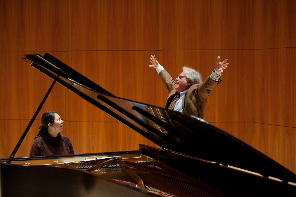 A musician stands triumphantly over a piano with his arms spread out, with another person sitting playing the piano