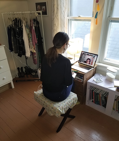 professor sitting at a small desk in a room that looks like an attic or hallway, with a student's face on a laptop in front of them.