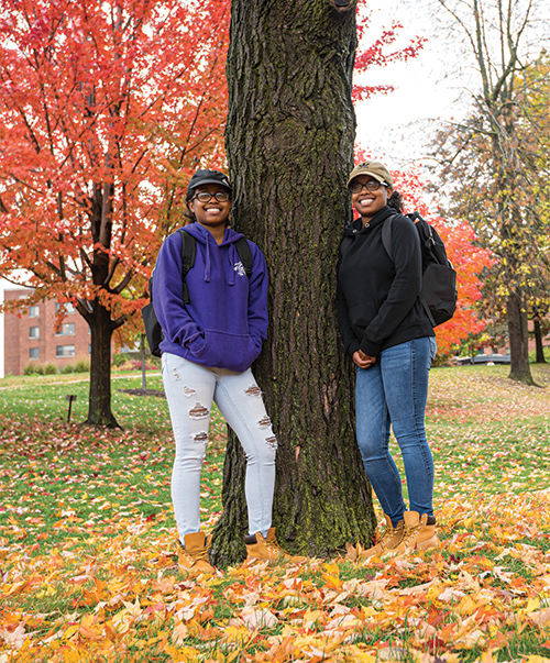 Destinee and Deziree Bell wearing Rochester sweatshirts and caps and carrying backpacks, posing under a fall tree.