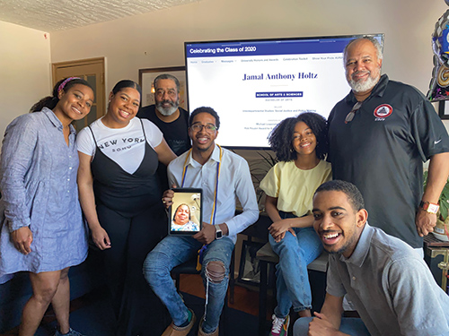 group photo of a famil shows a large screen behind them with the words CELEBRATING THE CLASS OF 2020: JAMAL ANTHONY HOLTZ while the family smile and hold an iPad that shows a woman's face on the screen.