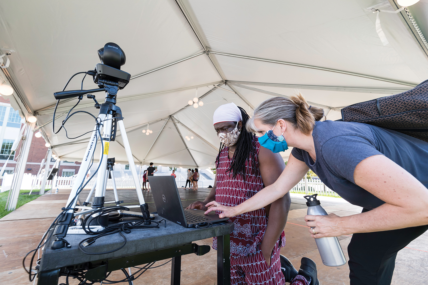 two instructors, both in masks, leaning over a laptop and camera in an outdoor tent as student dancers prepare in the background.