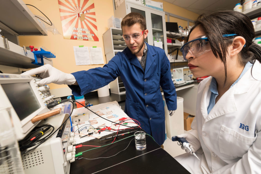 Two student researchers in a lab, wearing safety equipment and altering electronic panels