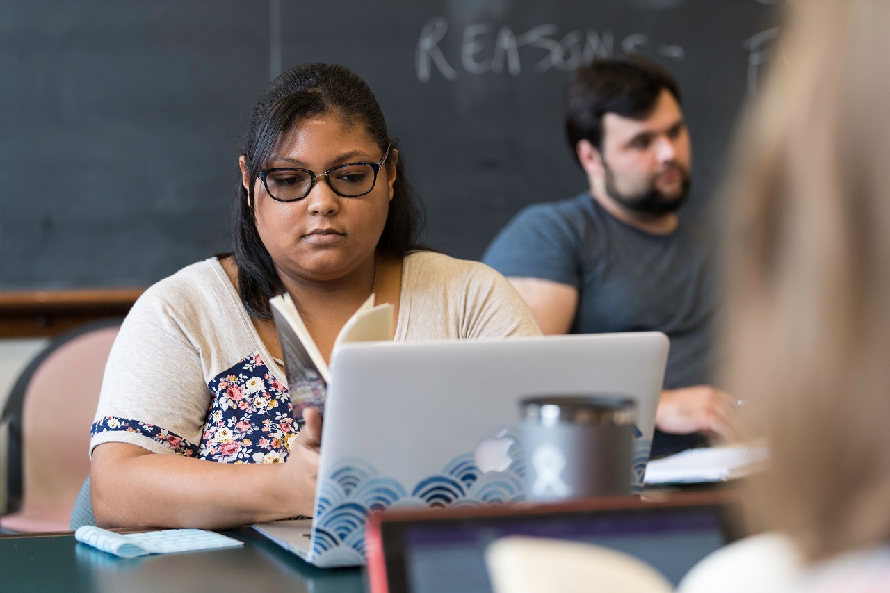 A University of Rochester student in a classroom, with an open book in hand and a laptop in front of her