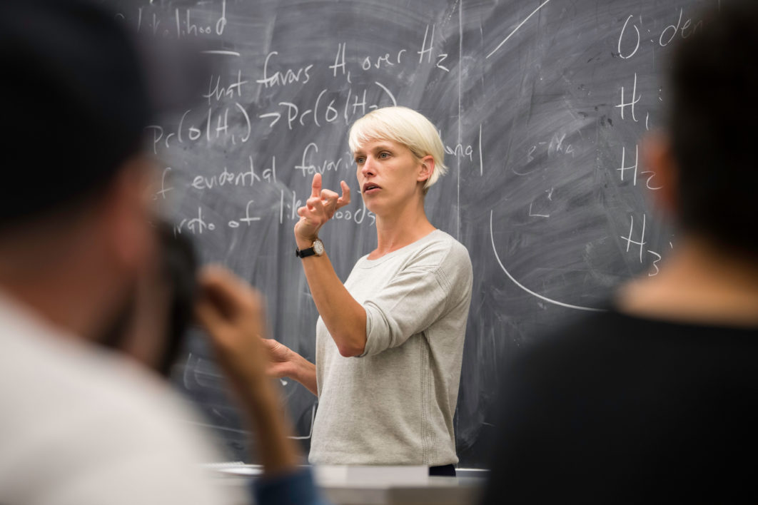 A professor in front of a chalkboard at the front of a classroom, speaking to her students
