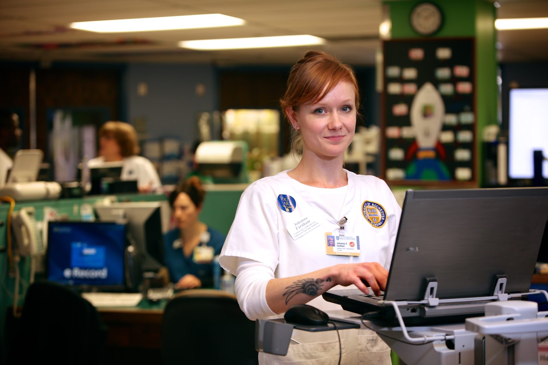A nursing student smiles for the camera in a hospital setting while at a computer