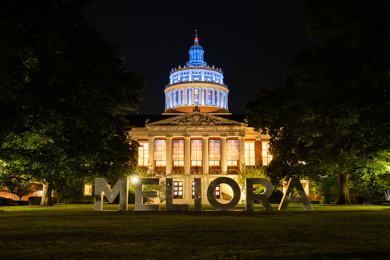 A view of Rush Rhees Library lit up at night with large letters spelling Melilla in the foreground.