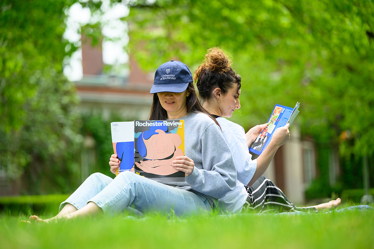 Two people with their backs together reading Rochester Review while sitting in the grass on the quad.