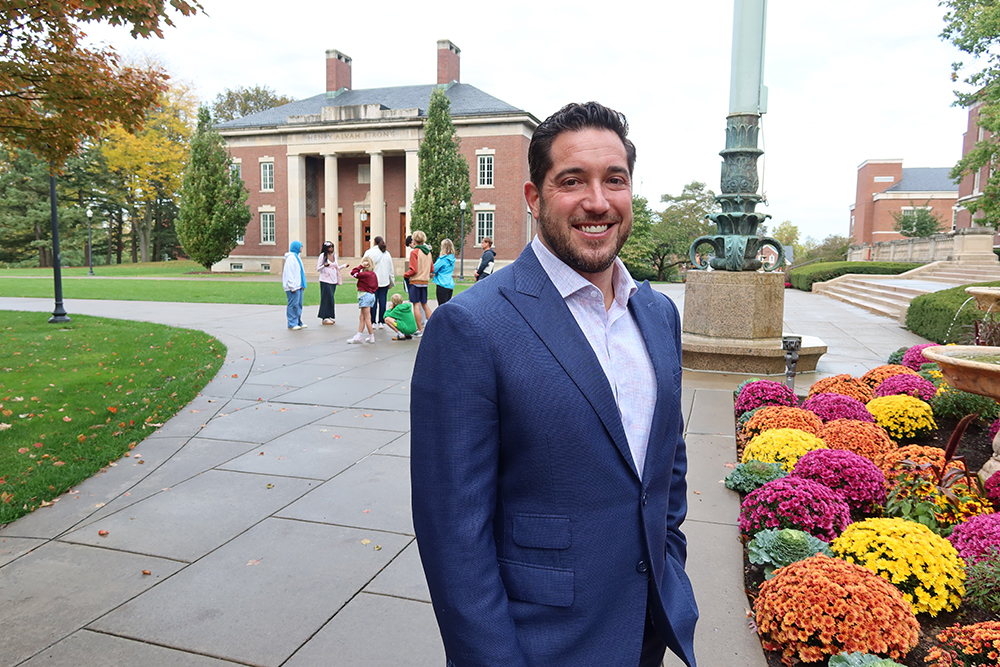 A person wearing a blue suit stands on a campus path with people and buildings in the background.