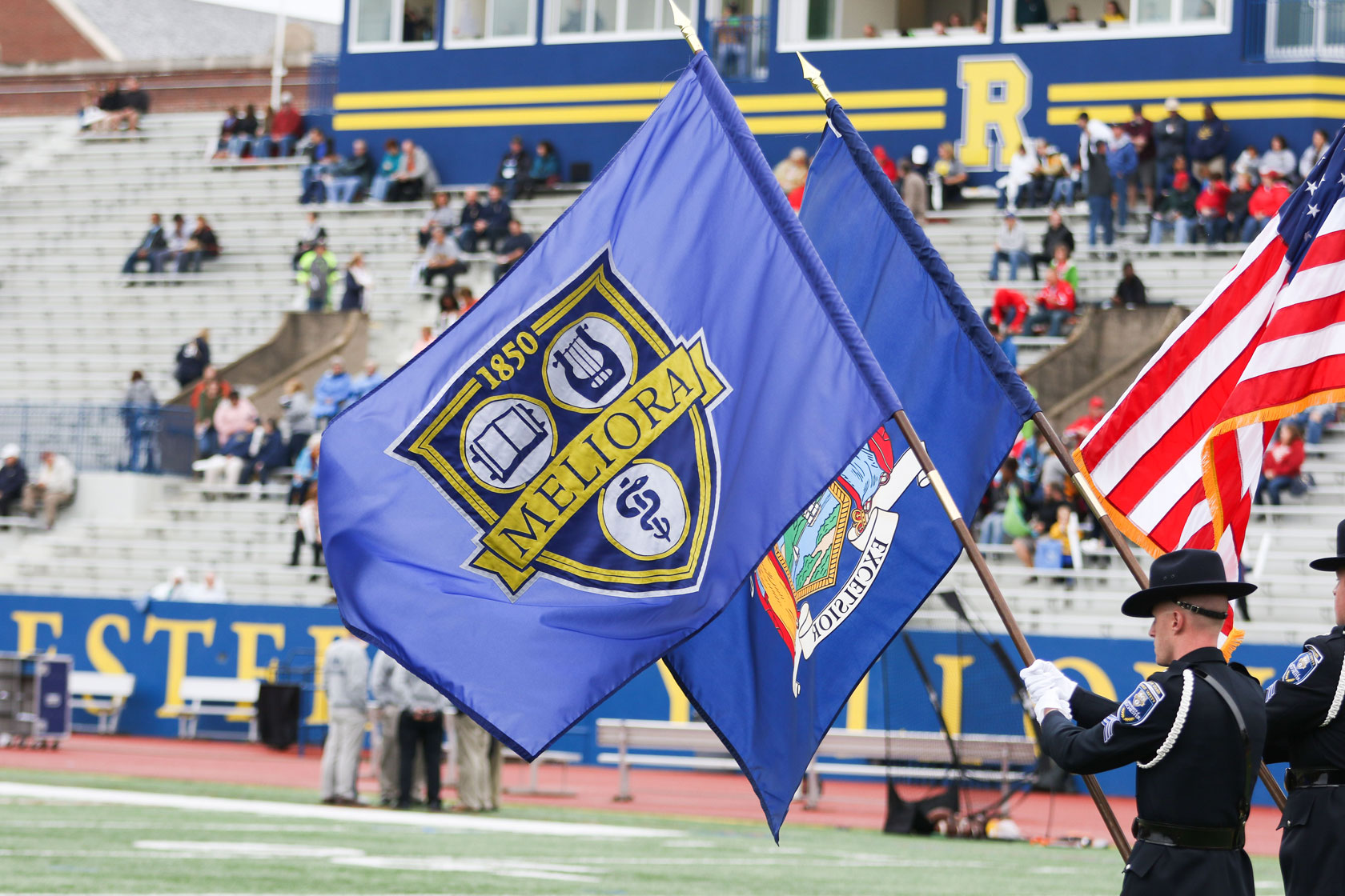 Flag bearers holding the University of Rochester flag, New York State flag, and American flag in a stadium.