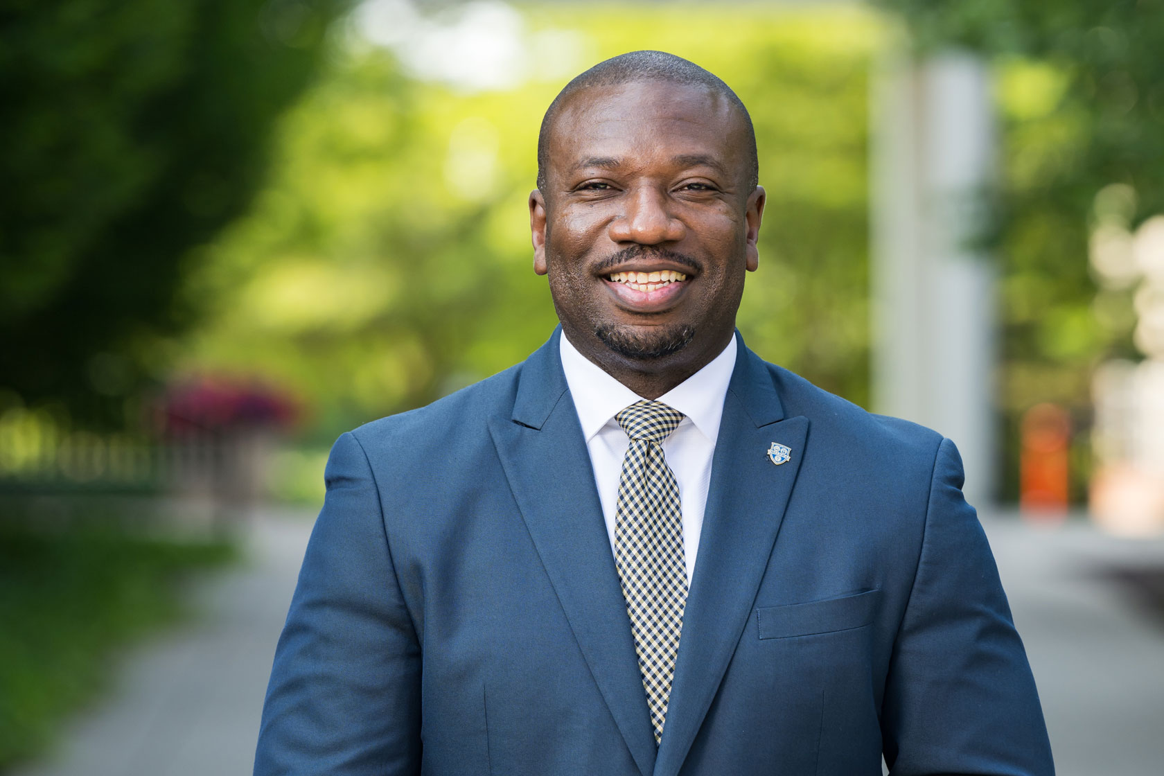 Quchee Collins, Associate Vice President of Public Safety and Chief Public Safety Officer, in a blue suit smiling outdoors with greenery in the background.