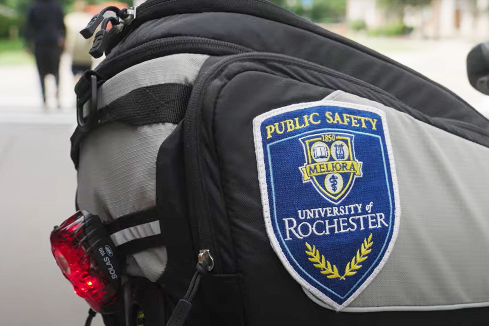 Feature image Close-up of a backpack with a "Public Safety, University of Rochester" patch and a red bike light.