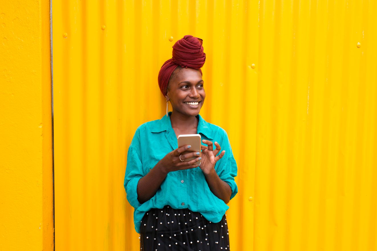 A young Black woman smiling on her phone, wearing a turquoise shirt and in front of a yellow wall