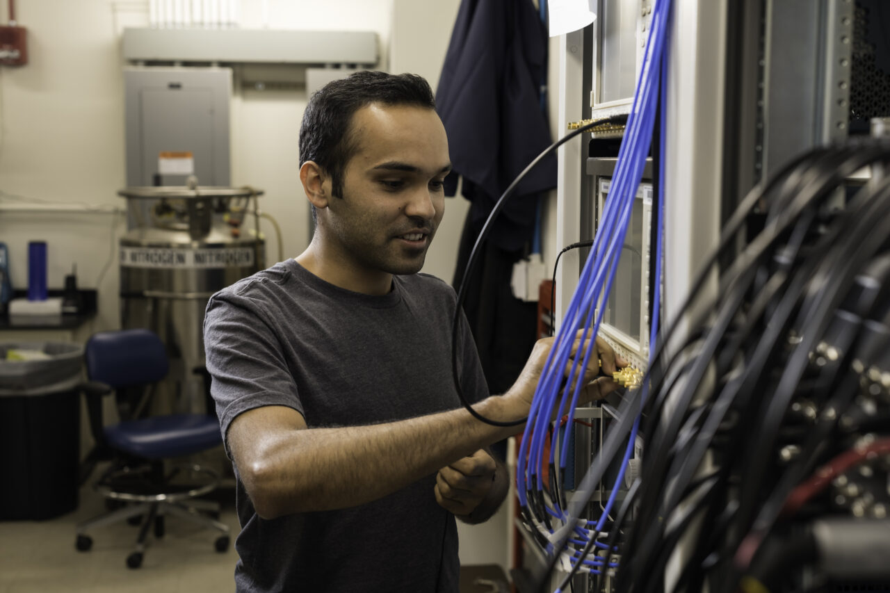 PhD student uses an Arbitrary Waveform Generator to manipulate qubits in a lab, pulling wires and working on machinery.