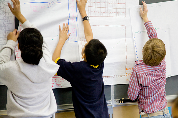 Three children stand in front of a classroom blackboard, reaching up to hang large sheets of paper displaying colorful graphs and charts. The charts include bar graphs, line graphs, and scatter plots. The children appear to be working together on a math or data project.