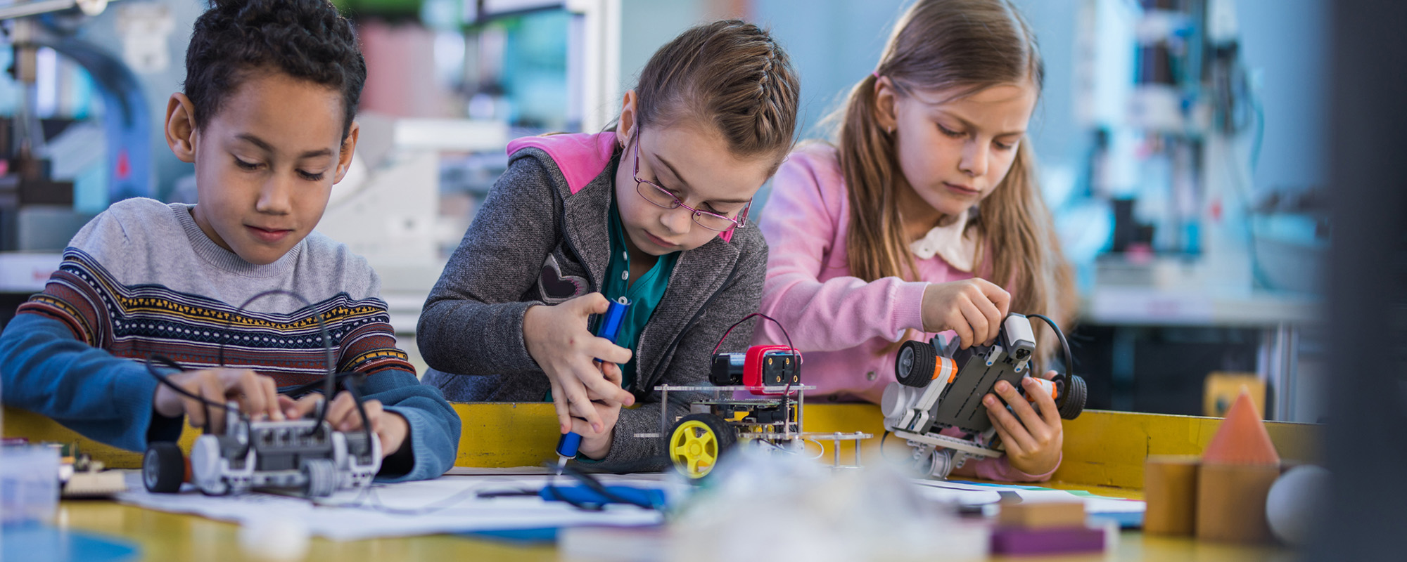 Three children work on small wheeled robots at a classroom table. Each child uses tools to assemble or adjust their robot. The background shows shelves and equipment, suggesting a STEM learning environment